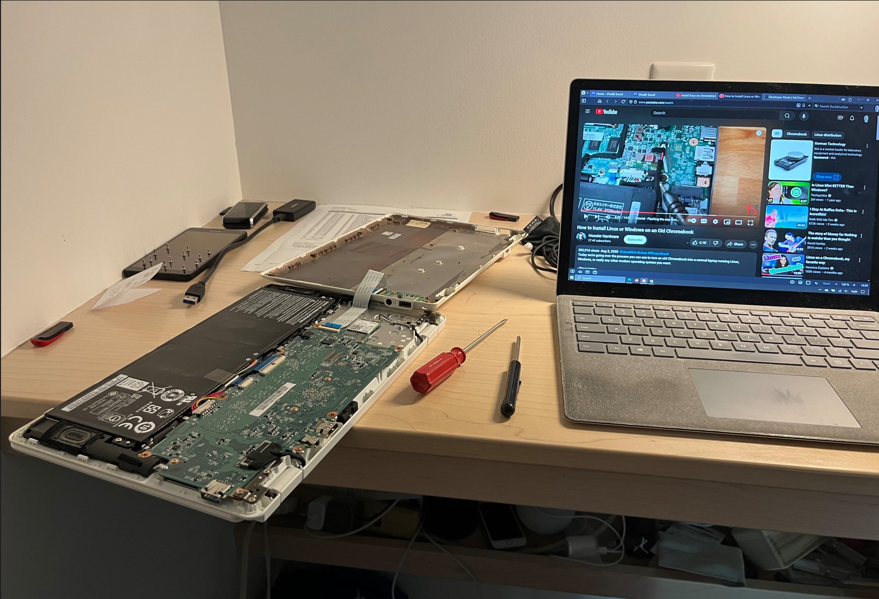 On a desktop, two laptops. The one on the right is open to a Youtube vide. The one on the left has its case open, showing the motherboard